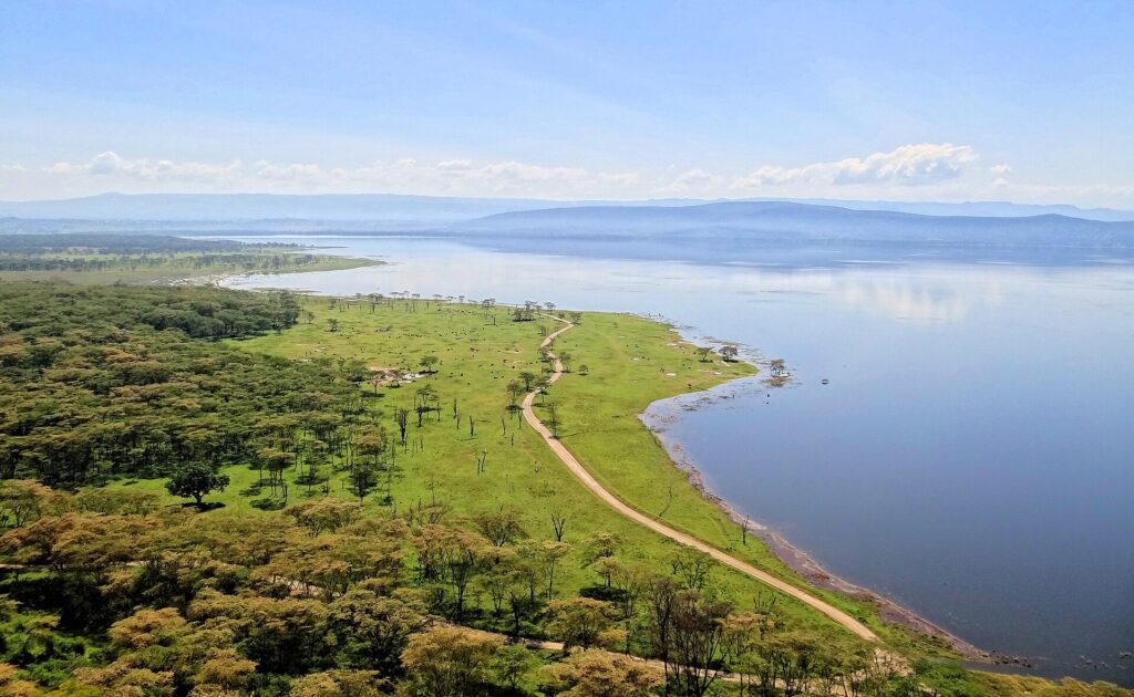 Pohled z Baboon Cliff dolů na břeh jezera, cesta podél zelené louky se zvířaty, akáciové háje a jezero až ke kopcům