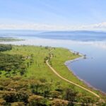 Pohled z Baboon Cliff dolů na břeh jezera, cesta podél zelené louky se zvířaty, akáciové háje a jezero až ke kopcům