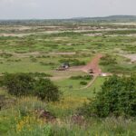 Výhled z Observation Hill na zelenou pláň se skvrnami soli, rudou cestu, tři vozy 4x4 zaparkované u malé okrové budovy a v dálce nízké kopce