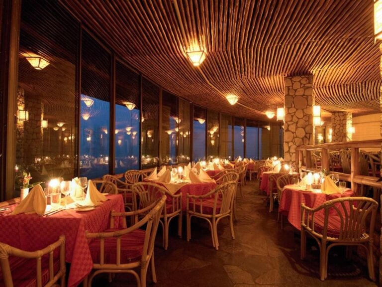 Jídelna restaurace osvětlená svíčkami, s červenými ubrusy a ratanovými křesly pod dřevěným stropem